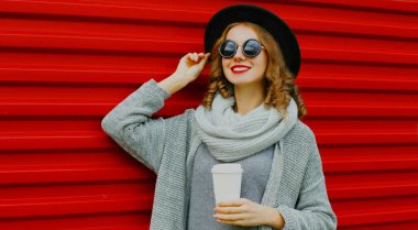 Elinde kahve fincanı olan güzel bir kadının portresi. Siyah yuvarlak bir şapka takıyor. Kırmızı arka planda gri örülü bir süveter.