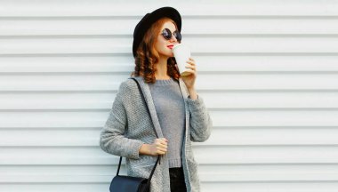 Elinde kahve fincanı olan, siyah yuvarlak şapkalı, beyaz arka planda gri örülü süveter giyen güzel bir kadının portresi.