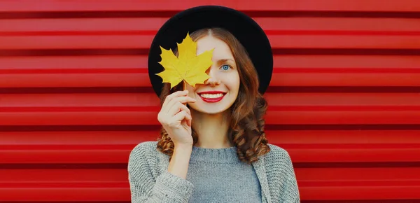 幸せの秋の肖像画若いです女性カバー彼女の目に黄色のカエデの葉を身に着けています黒丸帽子 グレーニットセーター上の赤の背景 — ストック写真