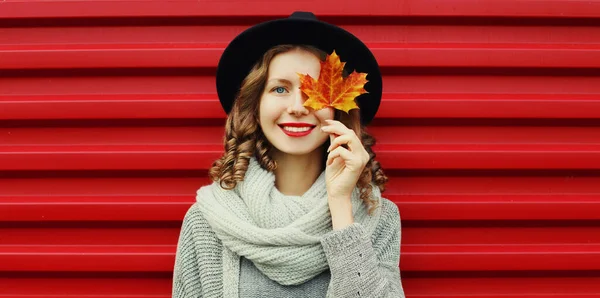 幸せの秋の肖像画若いです女性カバー彼女の目に黄色のカエデの葉を身に着けています黒丸帽子 グレーニットセーター上の赤の背景 — ストック写真