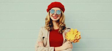 Sarı akçaağaç yaprakları ile gülen güzel, mutlu genç bir kadının sonbahar portresi gri arka planda kırmızı bir Fransız beresi takıyor.