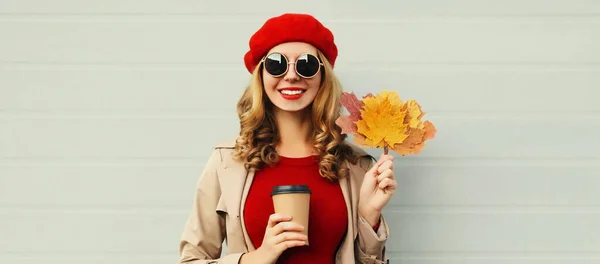 秋の肖像画の美しいです幸せな笑顔若いです女性とともに黄色のカエデの葉とカップのコーヒーを身に着けています灰色の背景に赤いフランス語ベレー帽 — ストック写真