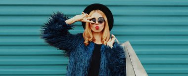 Şık bir sarışın kadın portresi. Alışveriş torbaları, mavi sahte kürk manto, renkli arka planda siyah yuvarlak şapka.