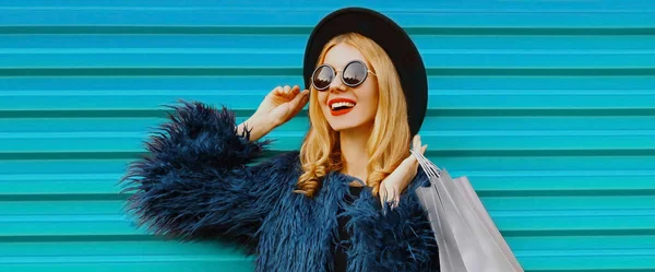 2つのショッピングバッグを身に着けているスタイリッシュなブロンドの女性のファッショナブルな肖像画青フェイクファーコート カラフルな背景に黒丸帽子 — ストック写真