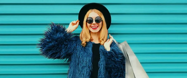 ファッショナブルな肖像画のスタイリッシュなブロンドの笑顔の女性でショッピングバッグを身に着けている青フェイクファーコート 黒丸帽子カラフルな背景 — ストック写真