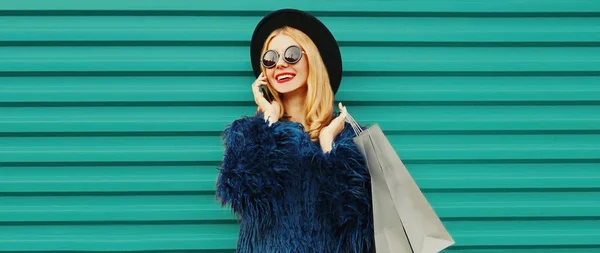青いフェイクファーのコートを着てスマートフォンを呼び出すショッピングバッグとスタイリッシュな笑顔の若い女性の肖像画 背景に黒丸帽子 — ストック写真