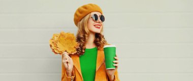 Sarı akçaağaç yaprakları ve kahve fincanı ile gülen güzel, mutlu genç bir kadının sonbahar portresi. Gri arka planda Fransız beresi takıyor.