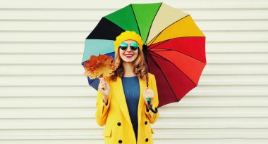Renkli şemsiyeli ve sarı akçaağaç yapraklı, beyaz arka planda palto ve bere takan mutlu, güler yüzlü genç kadının sonbahar portresi.