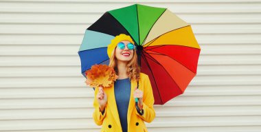 Renkli şemsiyeli ve sarı akçaağaç yapraklı, beyaz arka planda palto ve bere takan mutlu, güler yüzlü genç kadının sonbahar portresi.