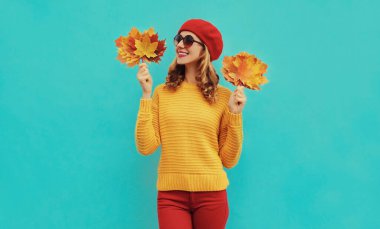 Sarı akçaağaç yapraklı mutlu genç bir kadının sonbahar portresi. Mavi arka planda kırmızı bereli örülü bir süveter giyiyor.