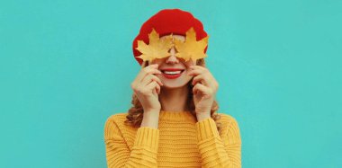 Sarı akçaağaç yapraklı mutlu genç bir kadının sonbahar portresi. Mavi arka planda kırmızı bereli örülü bir süveter giyiyor.