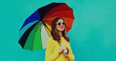 Mutlu, güler yüzlü, renkli şemsiyeli, mavi arka planda sarı örülü süveter giyen genç kadının sonbahar portresi.