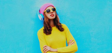 Kablosuz kulaklıkla müzik dinleyen, sarı örgü süveter ve mavi arka planda pembe şapka giyen modern genç bir kadının portresi.