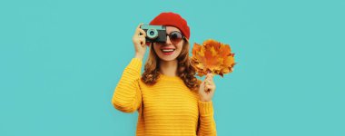 Film kamerası ve sarı akçaağaç yaprakları olan mutlu, genç bir kadının sonbahar portresi. Mavi arka planda Fransız beresi.