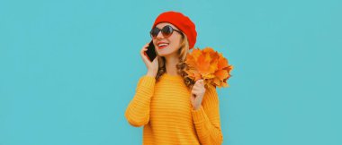 Sarı akçaağaç yapraklı mutlu genç bir kadının sonbahar portresi mavi arka planda kırmızı bir Fransız beresi takarak akıllı telefondan sesleniyor.