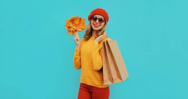 Alışveriş torbaları ve sarı akçaağaç yapraklarıyla mutlu bir genç kadının sonbahar portresi mavi arka planda bir süveter ve kırmızı bereyle