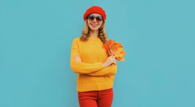 Sarı akçaağaç yapraklı mutlu genç bir kadının sonbahar portresi. Mavi arka planda kırmızı bereli örülü bir süveter giyiyor.