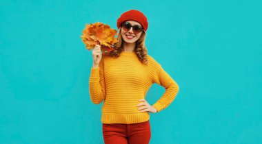 Sarı akçaağaç yapraklı mutlu genç bir kadının sonbahar portresi. Mavi arka planda kırmızı bereli örülü bir süveter giyiyor.