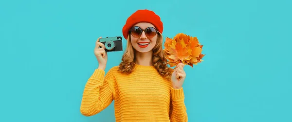 幸せな笑顔若い女性の秋の肖像画でフィルムカメラと黄色のカエデの葉はセーターを着て 青の背景にフランス語ベレー帽 — ストック写真