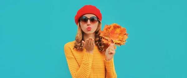 秋の肖像画の美しいです若いです女性とともに黄色カエデの葉吹いて彼女の唇を着てニットセーター 赤ベレー帽上の青の背景 — ストック写真