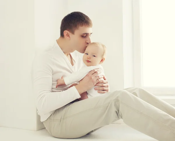 Jovem pai beijando bebê em casa na sala branca perto da janela — Fotografia de Stock