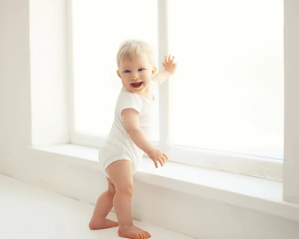 Sonriente bebé de pie en la habitación blanca en casa cerca de la ventana —  Fotos de Stock