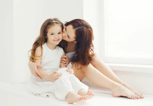 Madre cariñosa besando hija en casa en habitación blanca cerca de la ventana —  Fotos de Stock