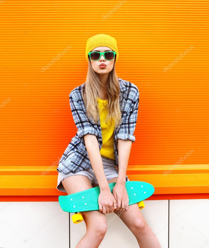 Fashion hipster cool girl in sunglasses and colorful clothes with skateboard having fun against the orange wall