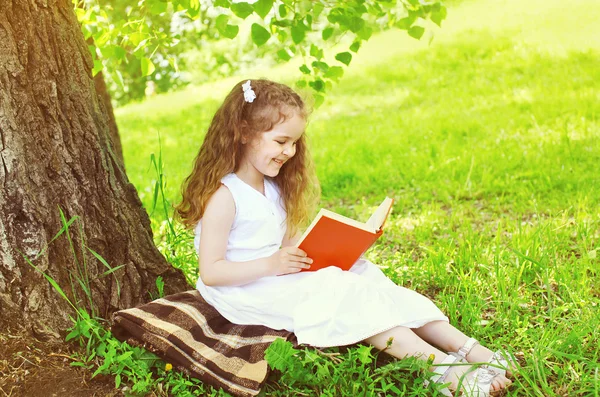 Улыбающаяся маленькая девочка читает книгу на траве возле дерева — стоковое фото