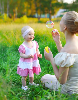 Mother and child playing blowing soap bubbles on the grass in su