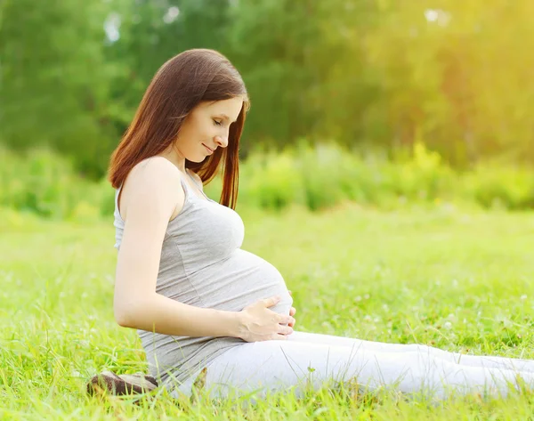 Portrait of happy pregnant woman sitting on the grass enjoys sun — Stock Fotó