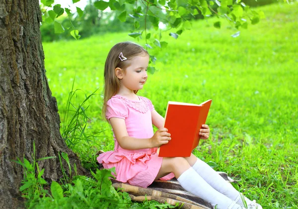 Маленькая девочка читает книгу на траве возле дерева в сумме — стоковое фото
