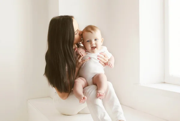 Mother kissing baby in white room at home near window — 스톡 사진