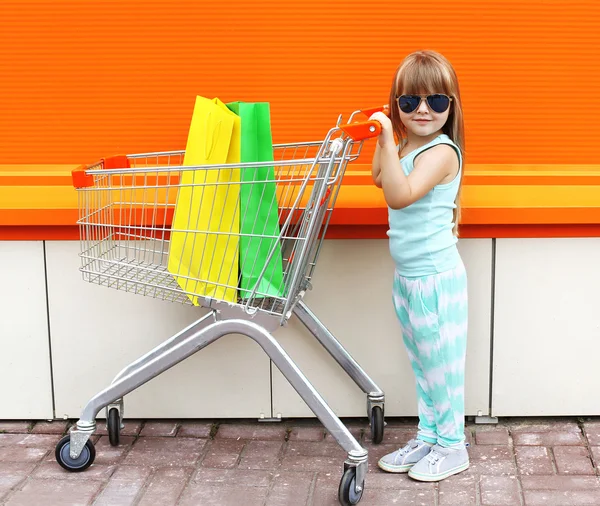 Little girl child and shopping cart with bags against the colorf — Zdjęcie stockowe