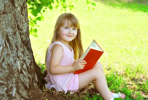 Little smiling girl child reading a book on the grass near tree — Stock Photo, Image