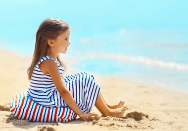 Little girl sitting on the sand beach near sea in summer day