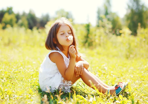 Niedliches kleines Mädchen, das Löwenzahnblume im sonnigen Sommer bläst — Stockfoto