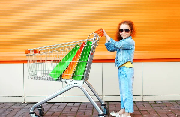 Criança menina feliz com carrinho de carrinho e compras coloridas — Fotografia de Stock
