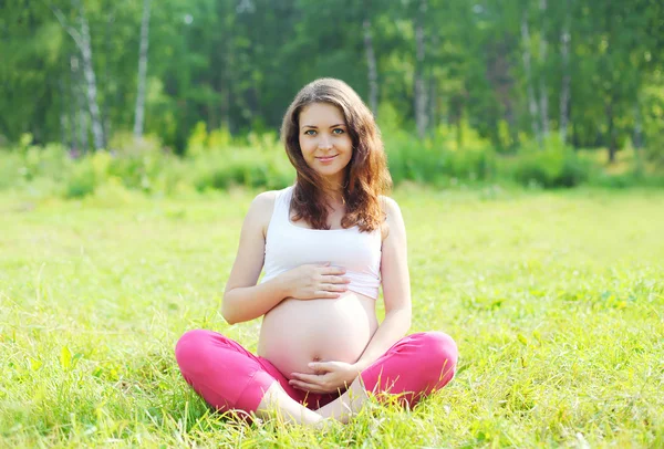 Happy young pregnant woman sitting on grass doing yoga in summer — 图库照片