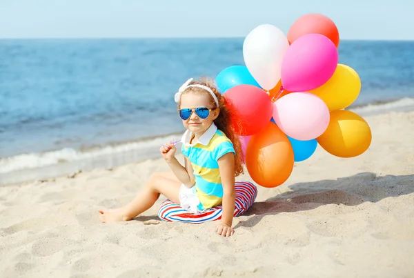 Niño feliz sentado en la playa de verano con globos de colores cerca —  Fotos de Stock