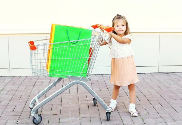 Criança menina feliz e carrinho de carrinho com compras coloridas — Fotografia de Stock