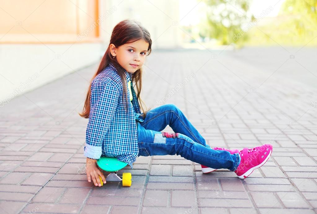 Fashion little girl child sitting on skateboard in city, wearing