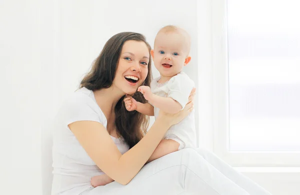 Feliz madre sonriente y bebé en casa en habitación blanca cerca de la ventana —  Fotos de Stock