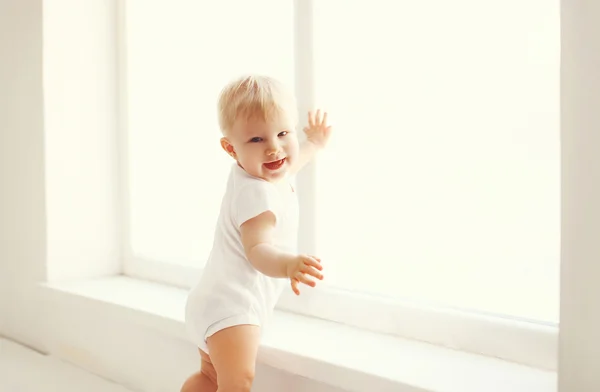 Pequeño bebé sonriente en habitación blanca en casa se para cerca de la ventana —  Fotos de Stock