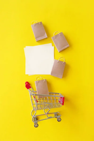 Mini trolley with small craft bags and blank white paper for lettering or shopping list on yellow background. The concept of shopping in stores and on the Internet.