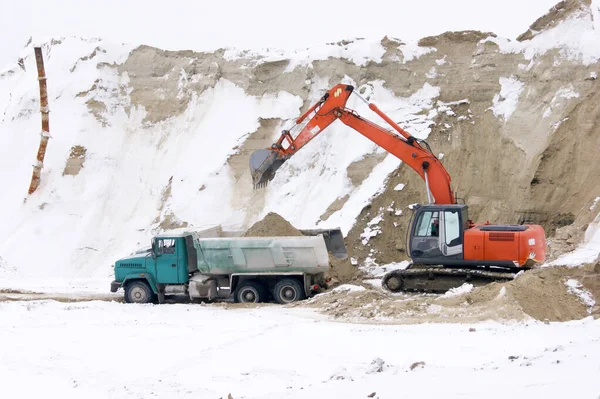 Crawler excavator in process of loading sand and soil to multi-ton mining truck. In the background there is a mountain of sand covered with snow.