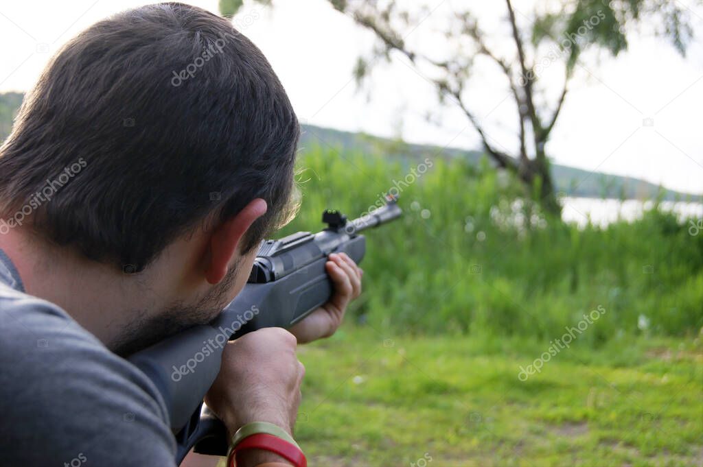 A young guy is preparing to shoot from a pneumatic weapon. Aims at a jar hanging from a tree.