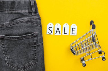 Düz yatış, siyah kot, beyaz etiketli alışveriş arabası ve sarı arka planda yazıt satışı. Alışveriş ve indirim konsepti. Kara Cuma.