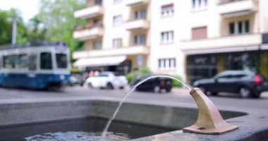İsviçre, Zürih 'te bir içme suyu kuyusu, tramvayların ve arabaların geçtiği ve insanların yürüdüğü.