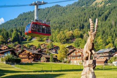 Wengen, İsviçre - 2 Ekim 2025: Mannlichen dağına giden kırmızı bir teleferik geleneksel İsviçre alp köyünün tebeşirlerinin üzerinde asılı duruyor